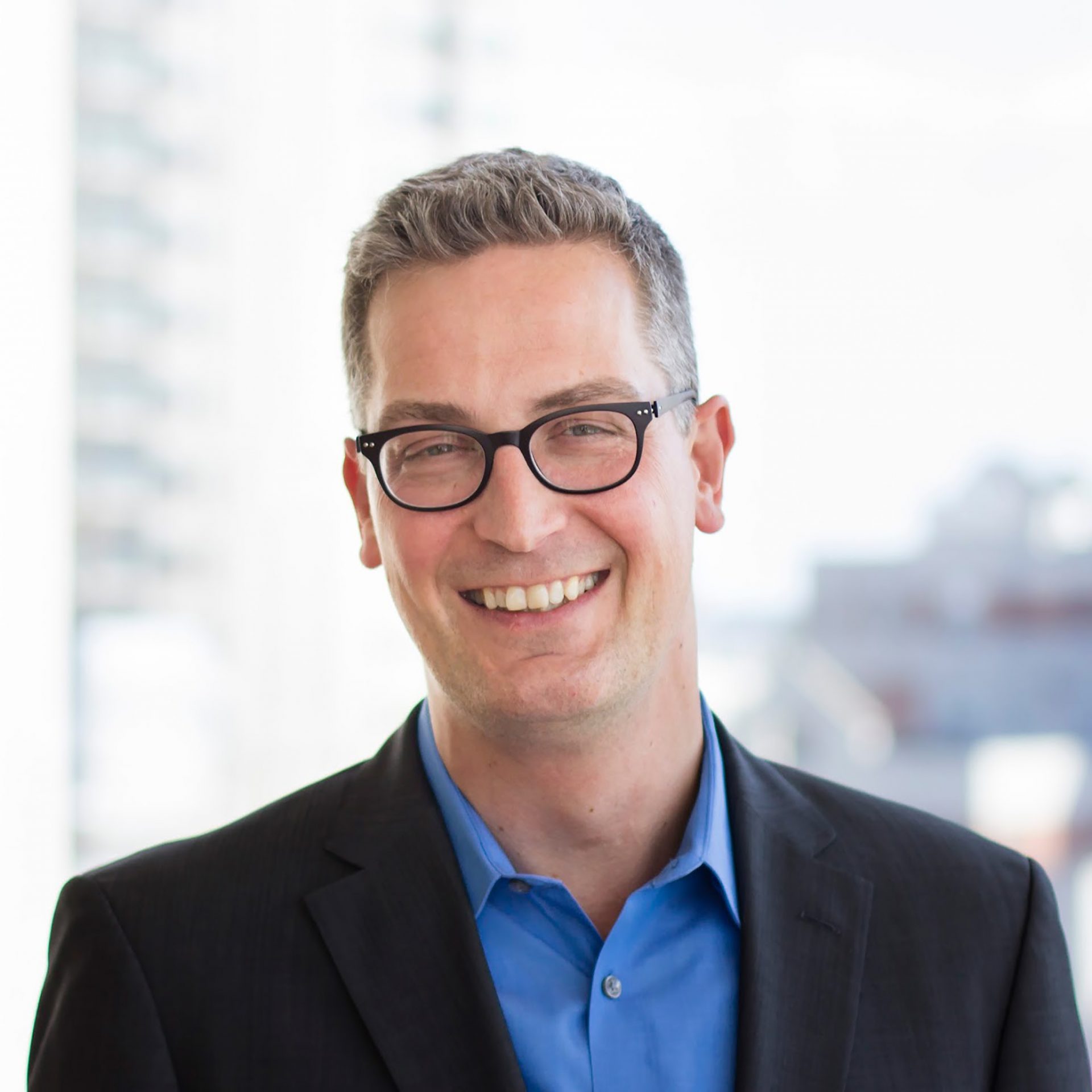 the photo shows a smiling Phil Buchanan, wearing glasses, a blue shirt, and a suit jacket..