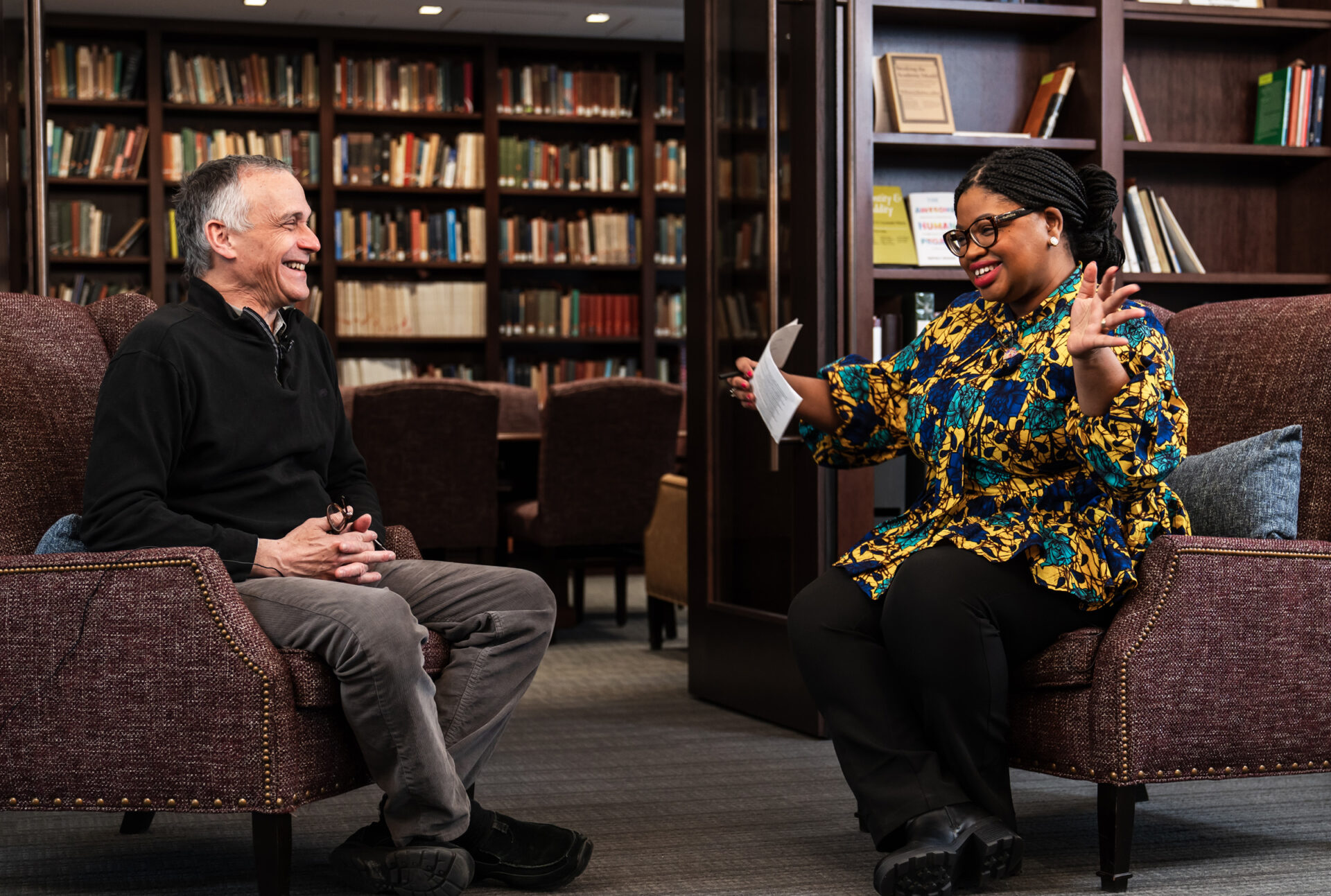 Two people sitting in library chairs having a conversation and smiling.